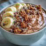 Two bowls of Chunky Monkey smoothie topped with granola, sliced banana, and chocolate chips on marble.