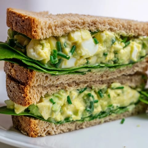Avocado Egg Salad Sandwich on whole grain bread with sliced tomato, served on a rustic wooden cutting board.