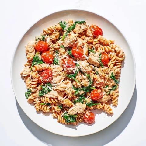 A steaming skillet of Greek Yogurt Chicken Pasta features tender chicken pieces, wilted spinach, and halved cherry tomatoes on a wooden table.